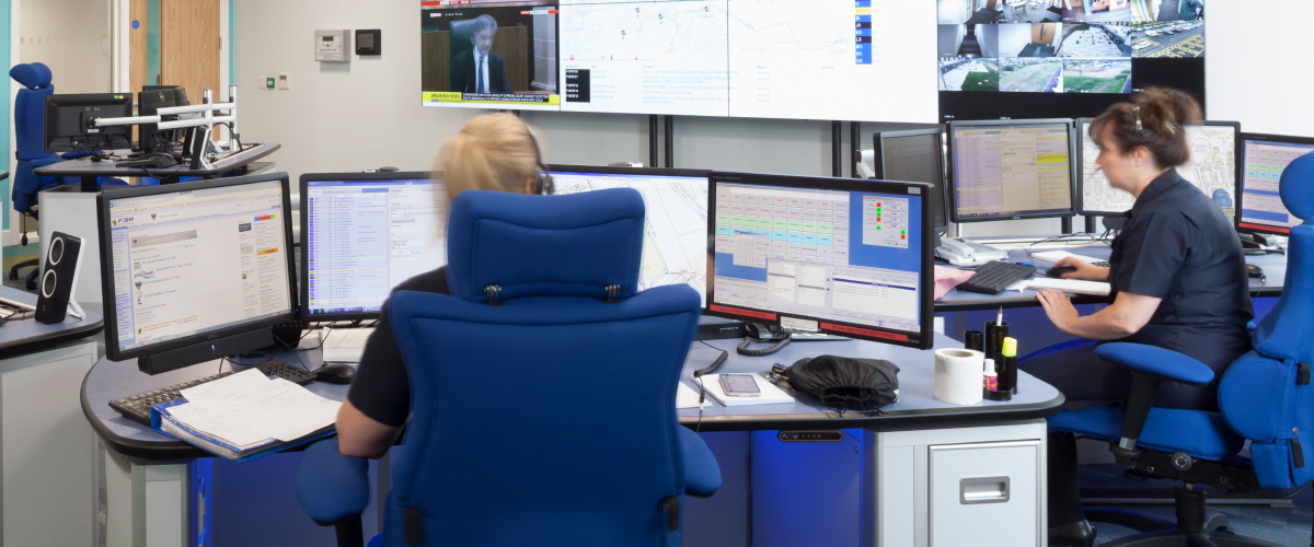 Two female control room operators working behind desks answering calls in the control room. Both wearing headsets with their backs to the camera.