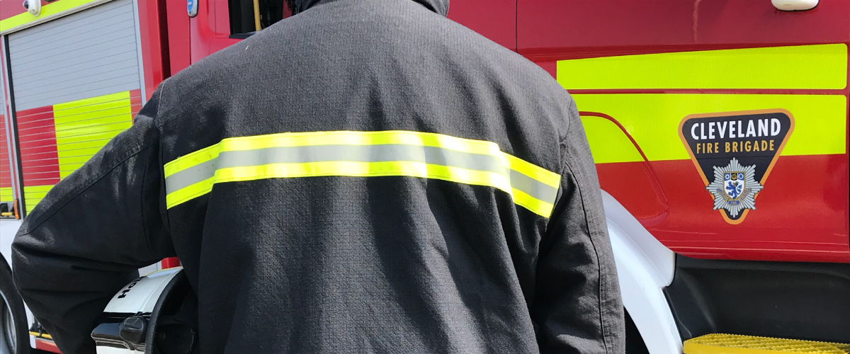 A firefighter in uniform facing towards one of our fire engines holding a white helmet
