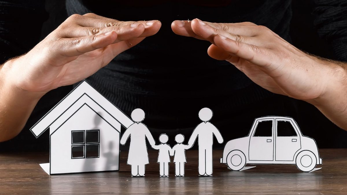 Paper cut outs of a house, car and family with a real pair of hands hovering over to protect