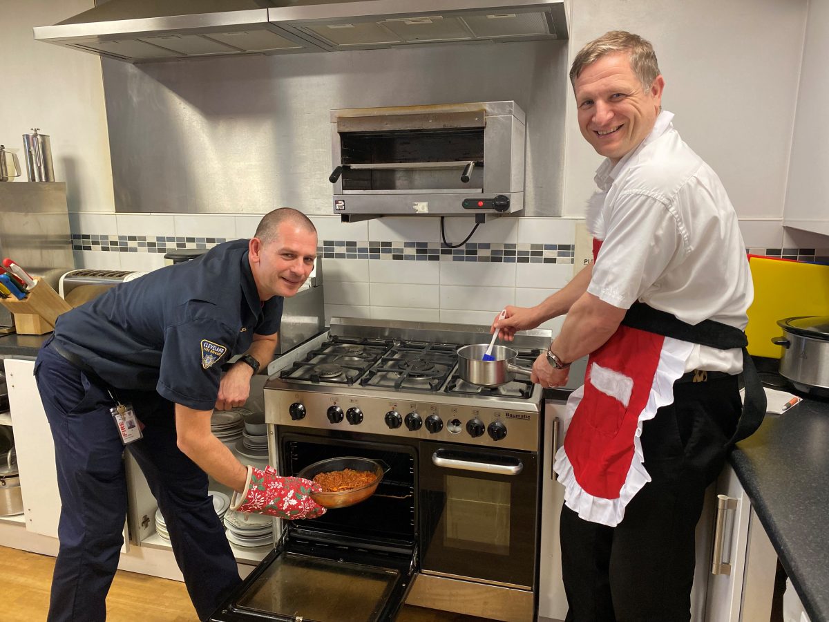 Two firefighters in a kitchen taking precaution when cooking. One has an apron on and the other has an oven glove. Both looking at camera and smiling.