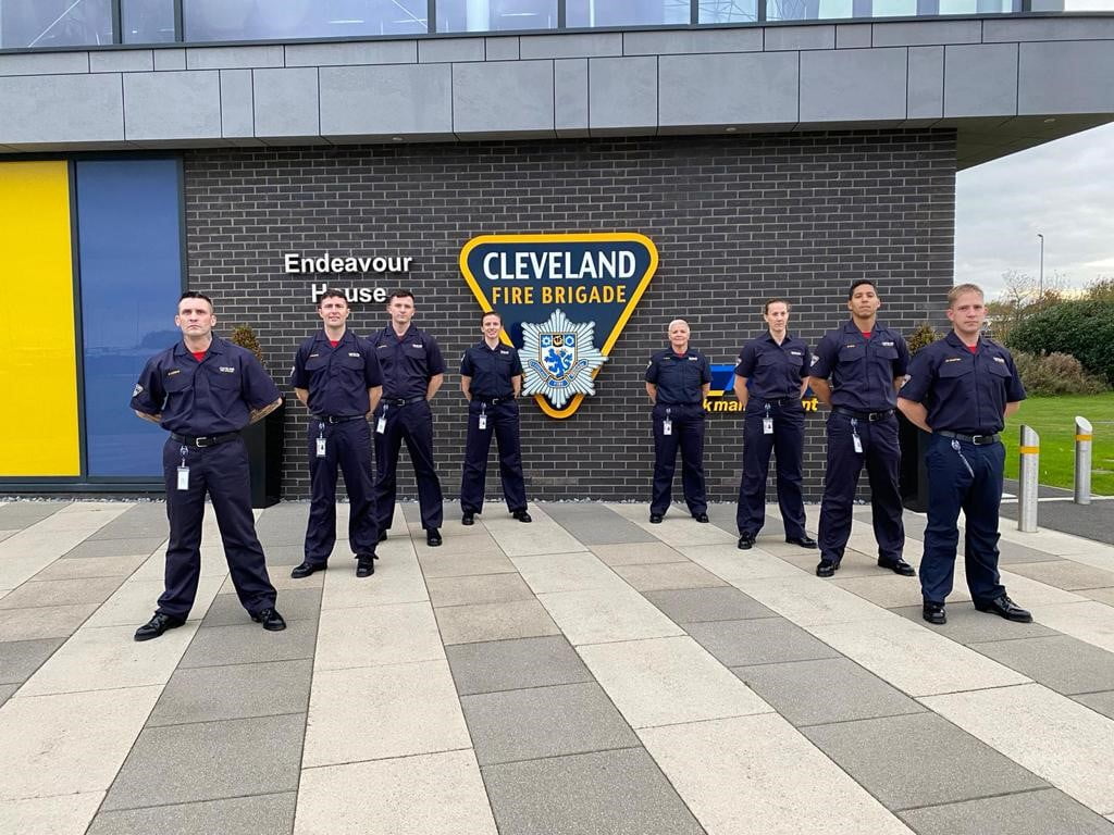 Picture of 8 on-call firefighters standing separately in uniform in front of HQ building.