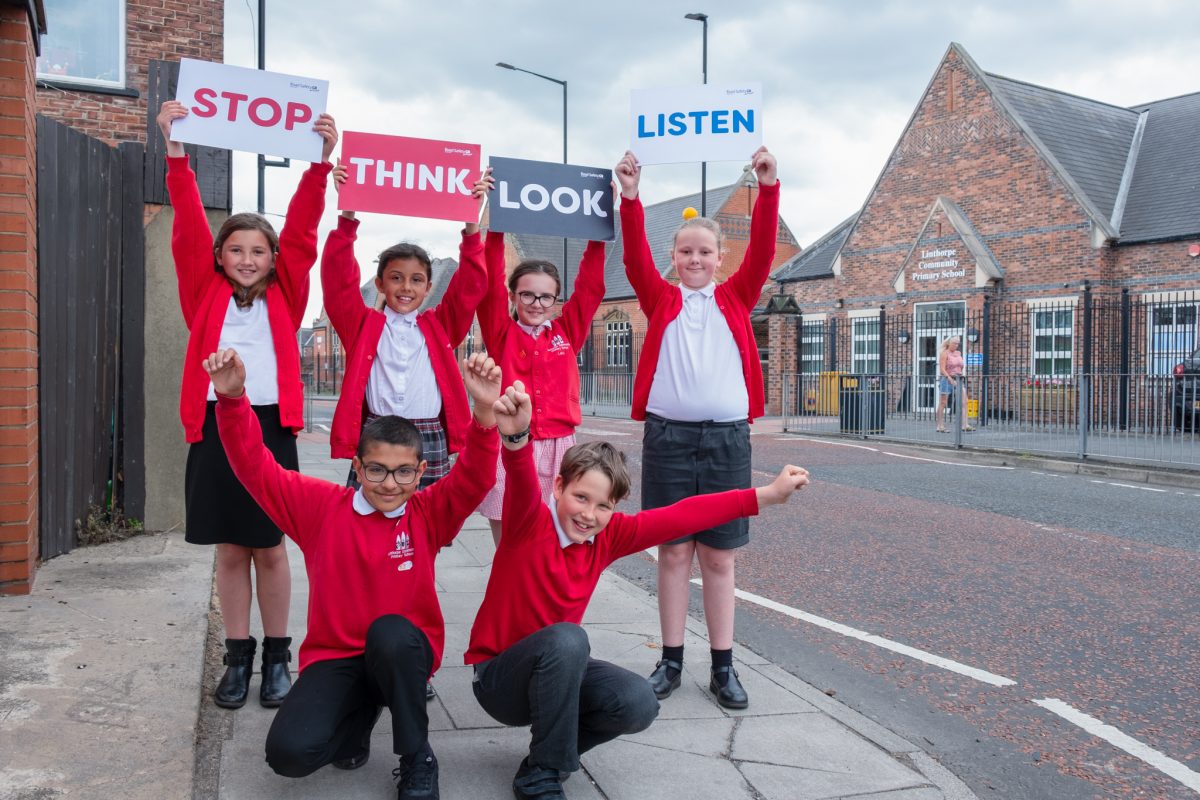 6 school children in red uniform outside of linthorpe primary school holding up road safety signs which read - stop, think, look and listen