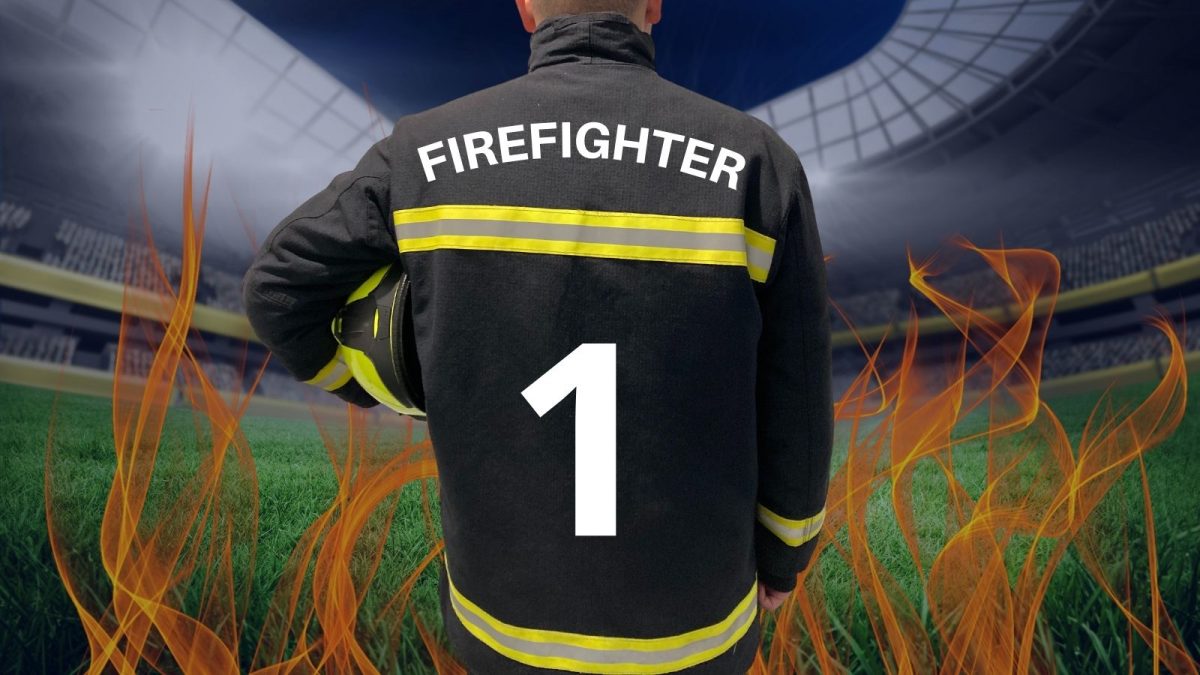 back of a firefighter in uniform holding an helmet looking out on to a football pitch