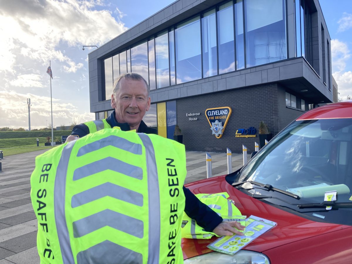 An image of a man holding up a reflective backpack and stood beside a red car