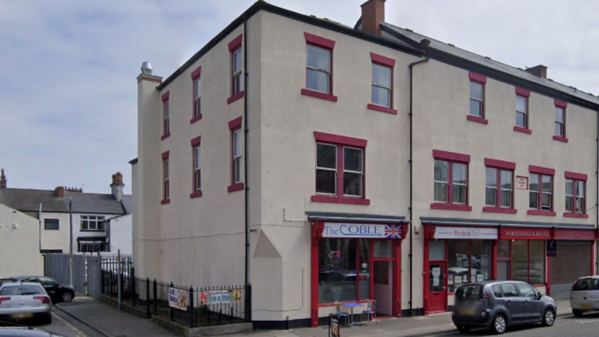 A white building containing shops with flats above with cars parked outside