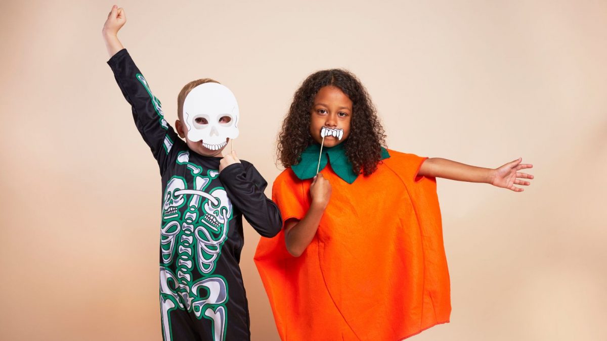 Two young children in fancy dress costumes. They are wearing a orange pumpkin costumer and a skeleton costume