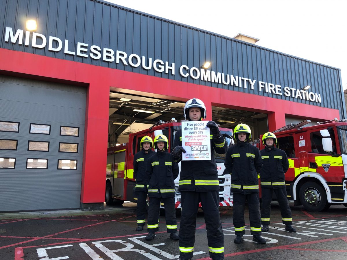 A group of firefighters stood outside of a fire station in a V formation holding a poster