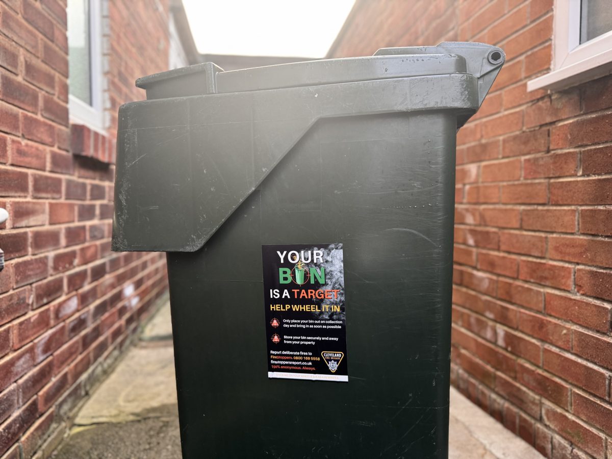 A green council waste bin in between two walls with a branded cleveland fire brigade sticker on the side to encourage residents to wheel it in