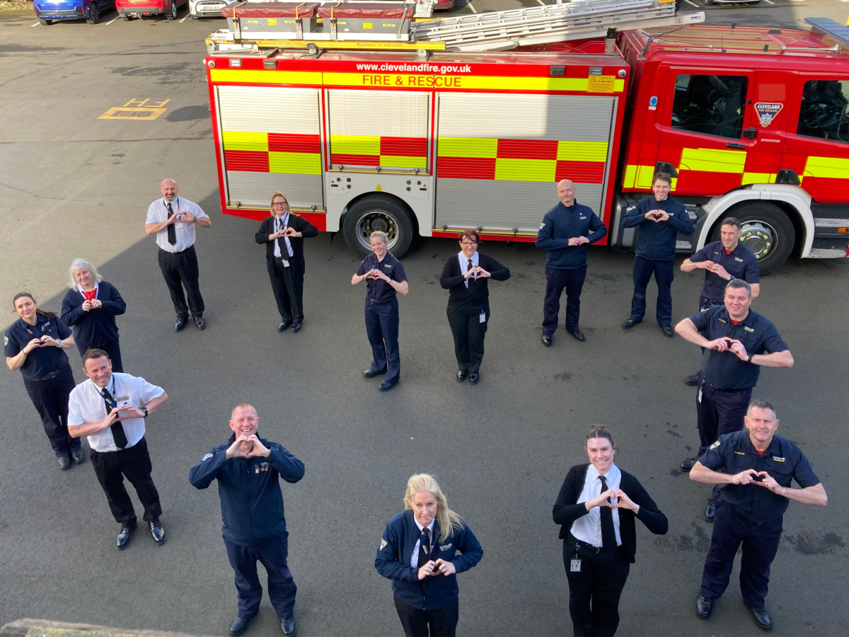 A group of male and female staff from the fire service stood in the formation of a heart and putting their hands in a heart shape. They are stood in front of a large red fire engine
