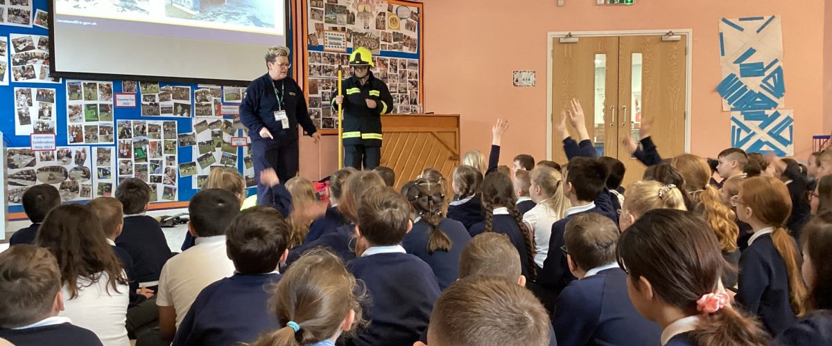 School Education Coordinator, Rita in uniform stood at the front of an assembly hall in front of a class of children. The children have their backs facing the camera and there is a firefighter dressed up in the corner.