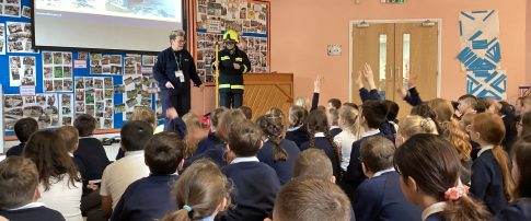 School Education Coordinator, Rita in uniform stood at the front of an assembly hall in front of a class of children. The children have their backs facing the camera and there is a firefighter dressed up in the corner.