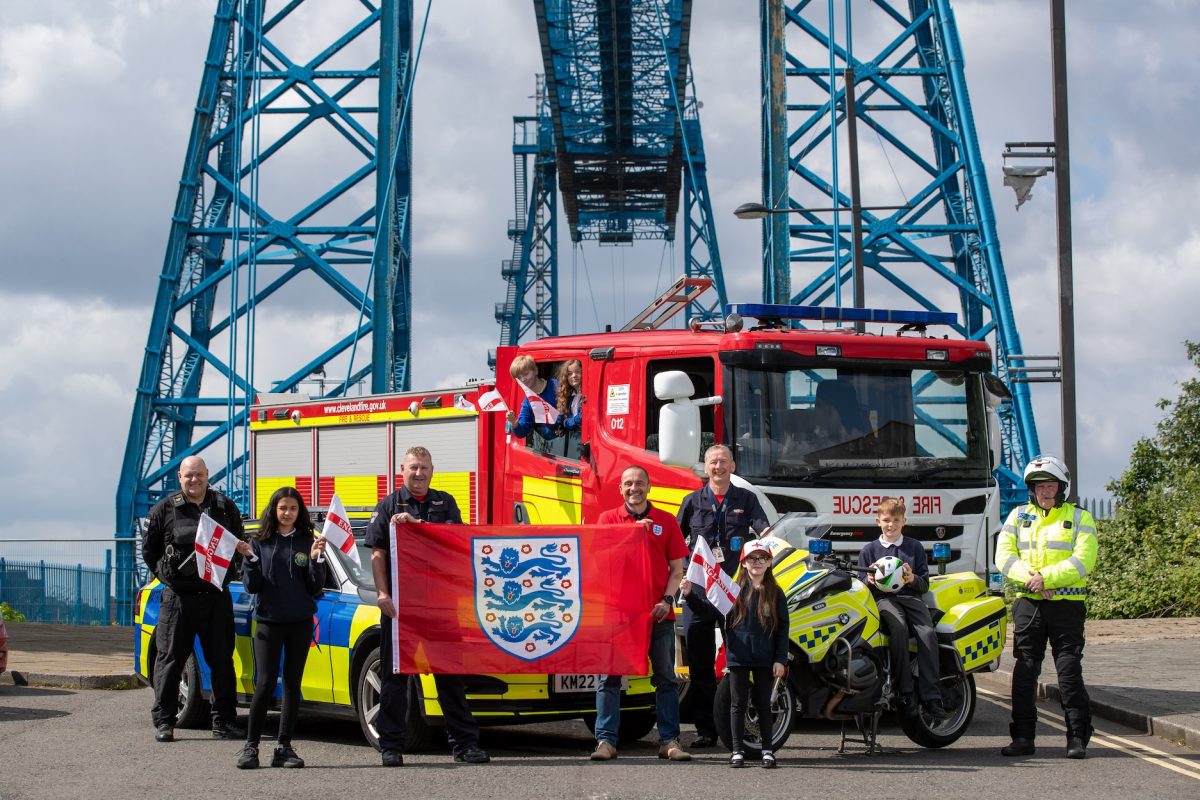 A group of children and adults stood on a blue bridge holding up a red England flag. Behind them is a red fire engine.