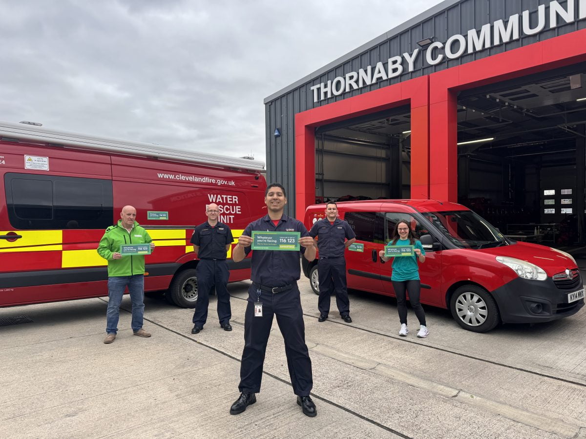 Five people stood in a triangle formation in front of a fire station and two red cars holding up a sign with the phone number for the Samaritans