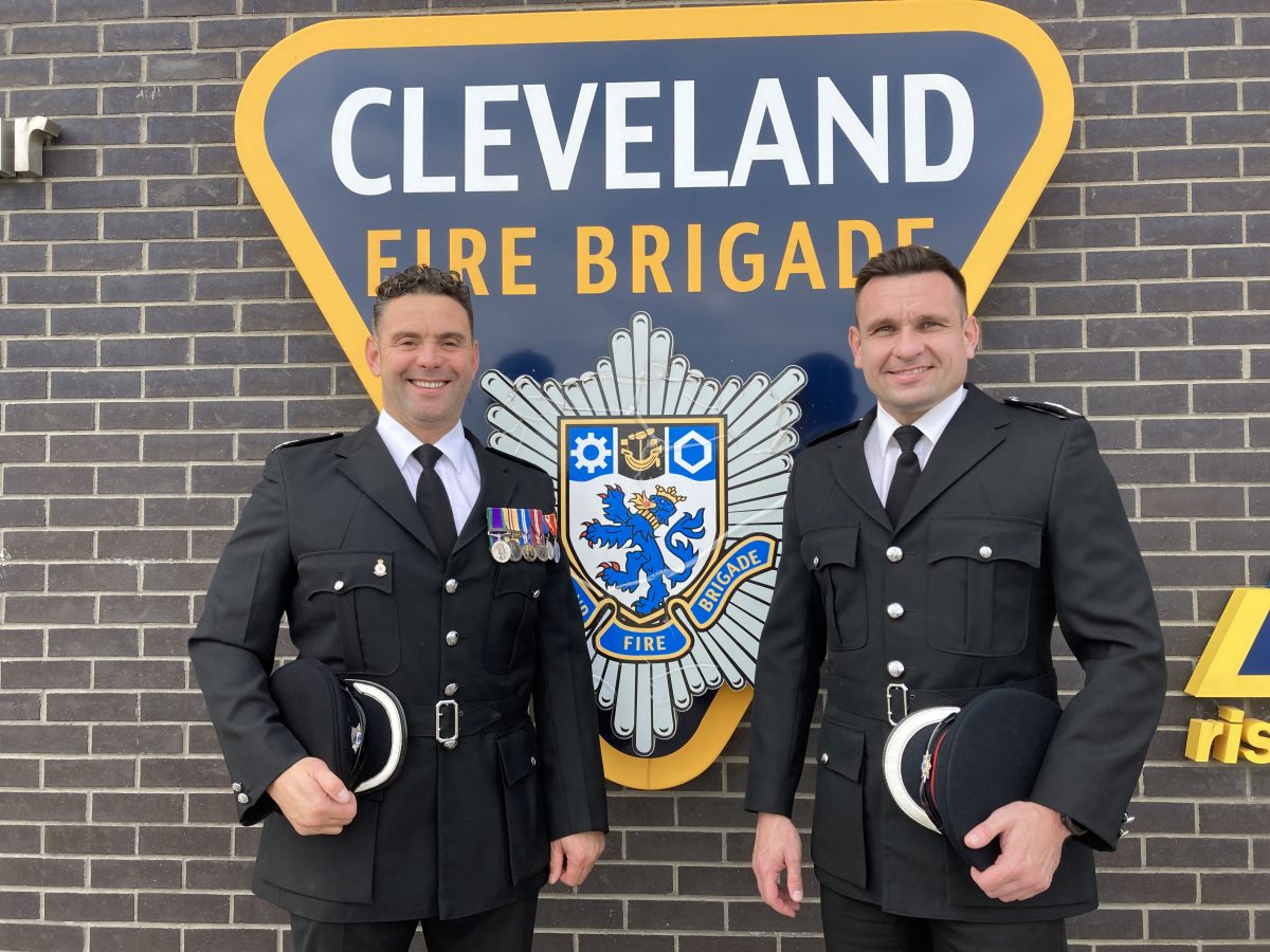Two male fire brigade personell in black uniforms stood in front of the blue and yellow Cleveland Fire Brigade badge.