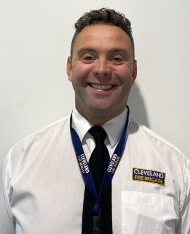 A portrait headshot of Lee Brown smiling wearing white shirt, black tie and a lanyard