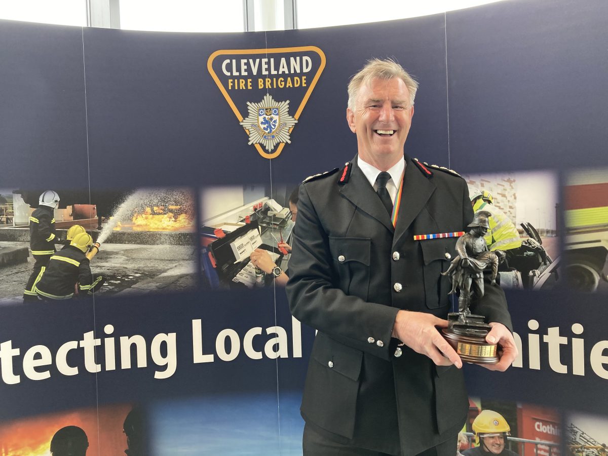 The Chief Fire Officer in his full dress holding his award in front of a stand that says protecting local communities