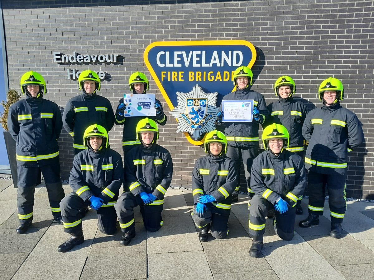 A picture of 10 trainee Cleveland Fire Brigade Firefighters in front of a Cleveland Fire Brigade Logo. Two of the trainee Firefighters are holding signs that say "I'm supporting Befriending Week" and "I'm supporting Befriending Week because it brings our community together"