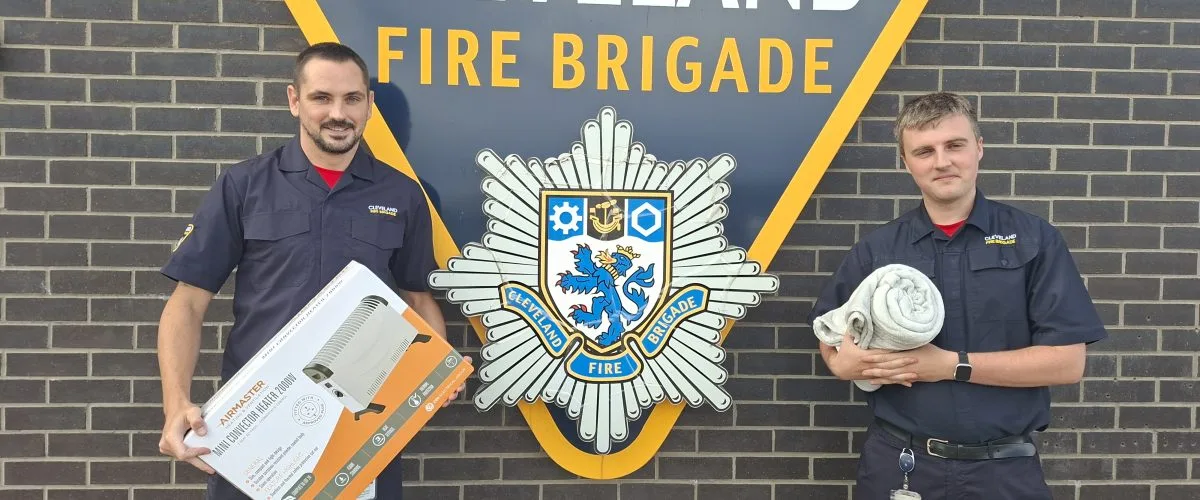 Two members of staff one with a heater and the other with a blanket in front of a cleveland fire brigade logo.