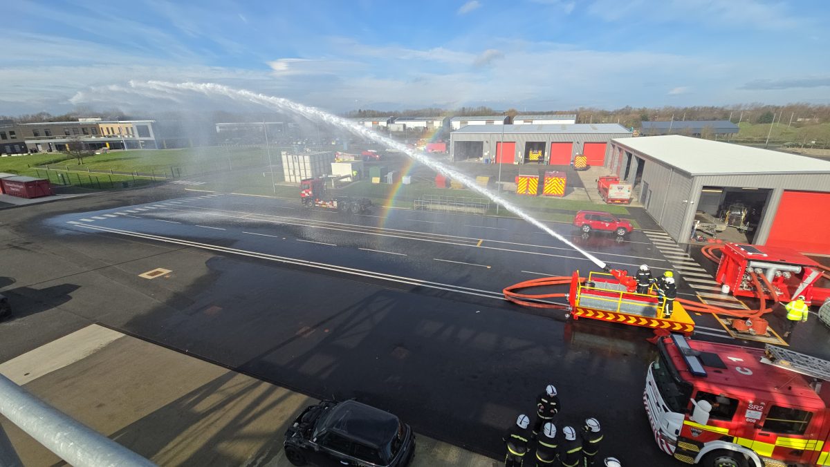 A landscape photo of a bright sunny day with blue skies that is overlooking Cleveland Fire Brigade headquarters in Hartlepool, North-East England. The image features a fire engine and a foam cannon, alongside multiple firefighters, with the cannon being used to shoot water into the sky. 