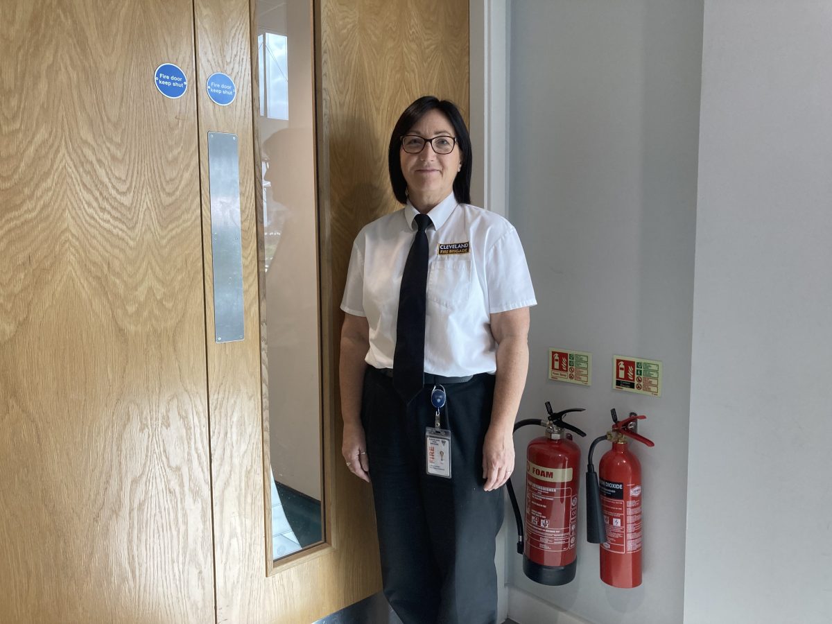 A picture of a woman in a white shirt and black trousers and tie stood next to fire doors and two fire extinguishers